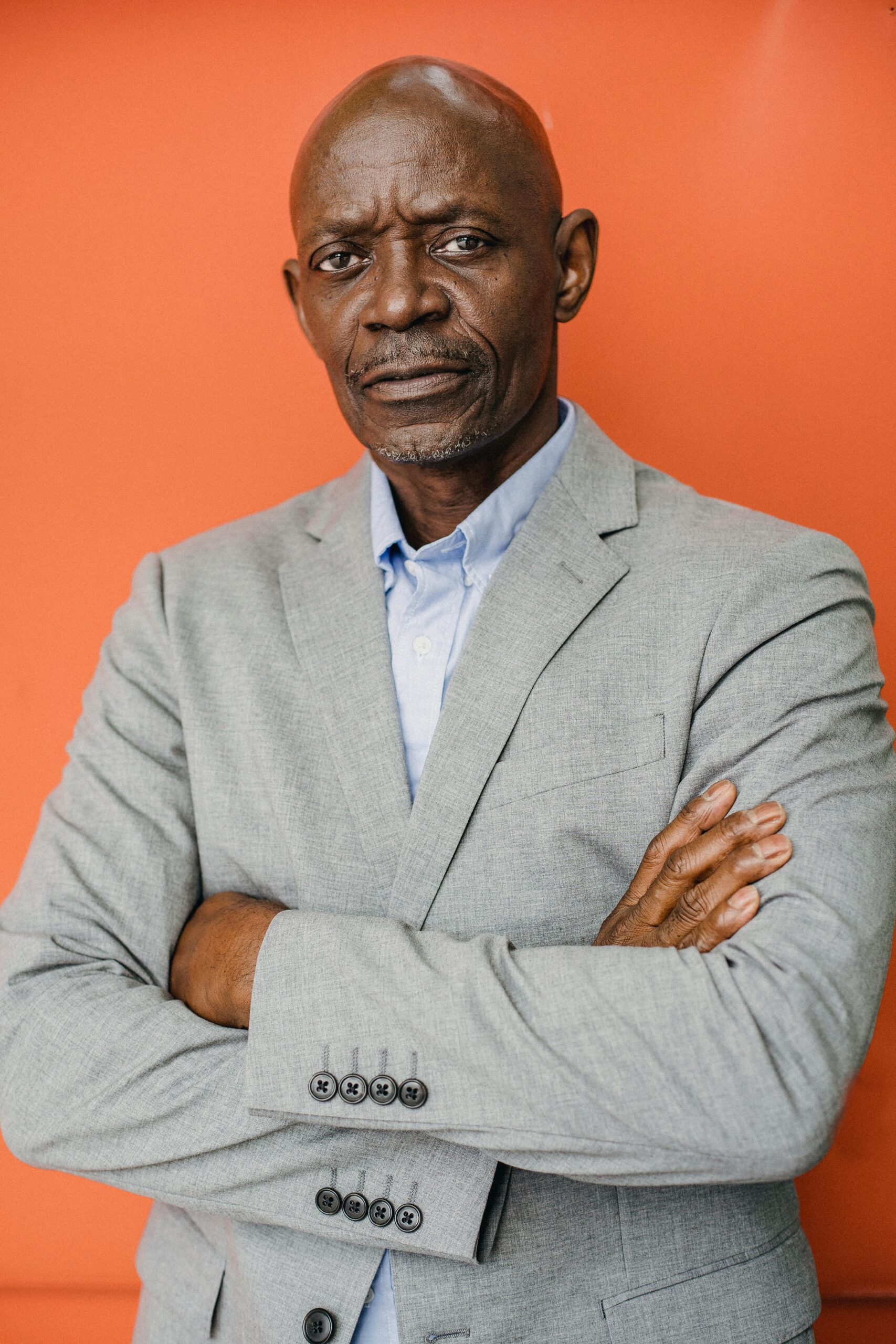 A serious African American man poses confidently in a grey suit against a vibrant orange backdrop.